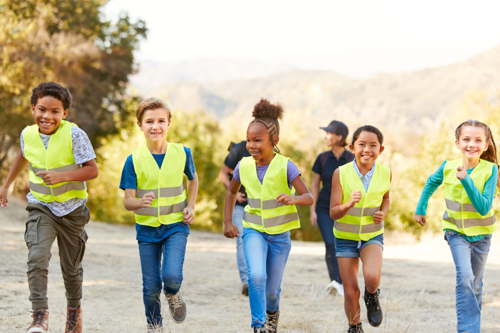 Children wearing reflective vests running outdoors on a sunny day.
