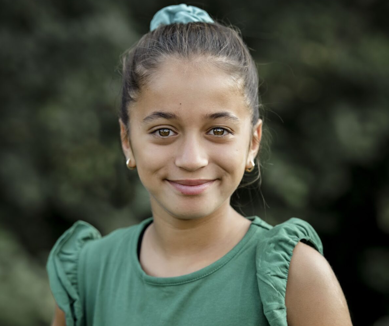 A young girl with a green top and a blue hair tie smiles gently.